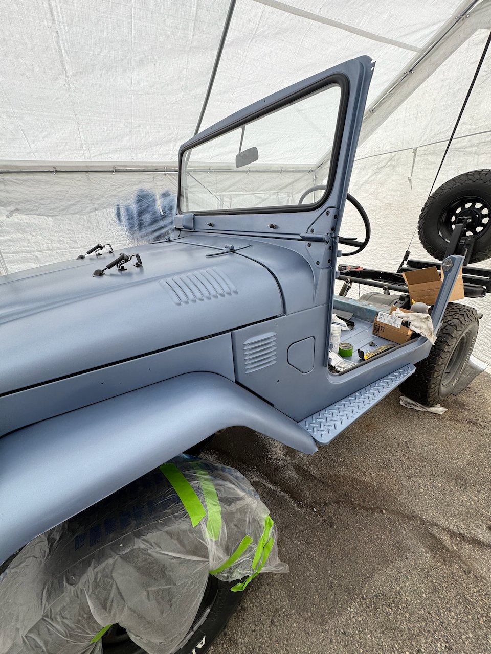 The FJ40 hood and side panels in fresh gray-blue paint, windshield folded up, rest of the truck still in pieces behind it